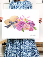 Load image into Gallery viewer, Woman holding a picture of tan sunhat with black bow beside a bunch of pink, coral, fuscia, and purple dahlias with green leaves
