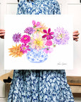 woman holding picture of Blue and White vase filled with bright dahlia flowers of different varieties. Colors used: purple, lilac, magenta, pink, yellow, royal blue, light blue, orange