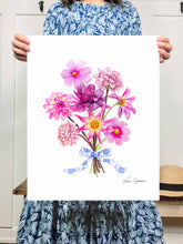 Load image into Gallery viewer, Woman in blue patterned dress holding a picture of Dahlias and cosmos in a bouquet tied together with a blue and white bow. colors featured are purples, fuscia, dusty rose, yellows, and blue and white
